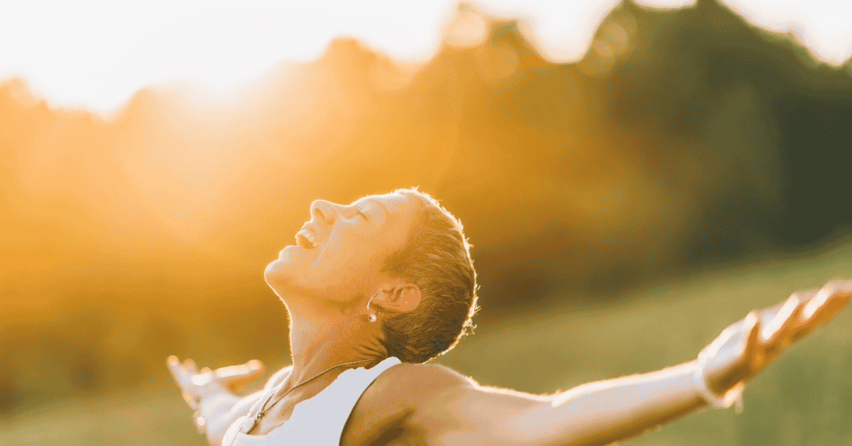 woman standing outside in a field withher arms stretched out to her sides and her head thrown back with a giant smile while being bathed in sunlight to symbolize letting go of expectations and moving into trust