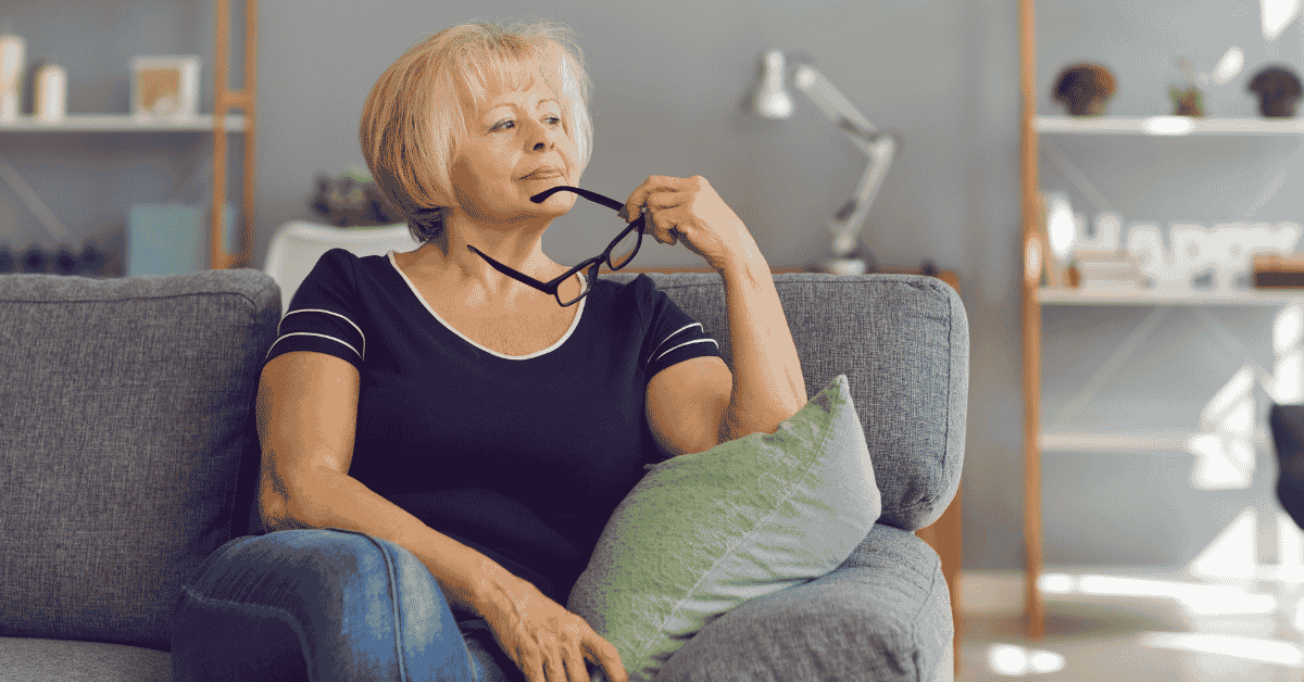 A woman sitting on a sofa with her eye glasses resting on her chin looking curious to represent going within to help heal old patterns as they arise