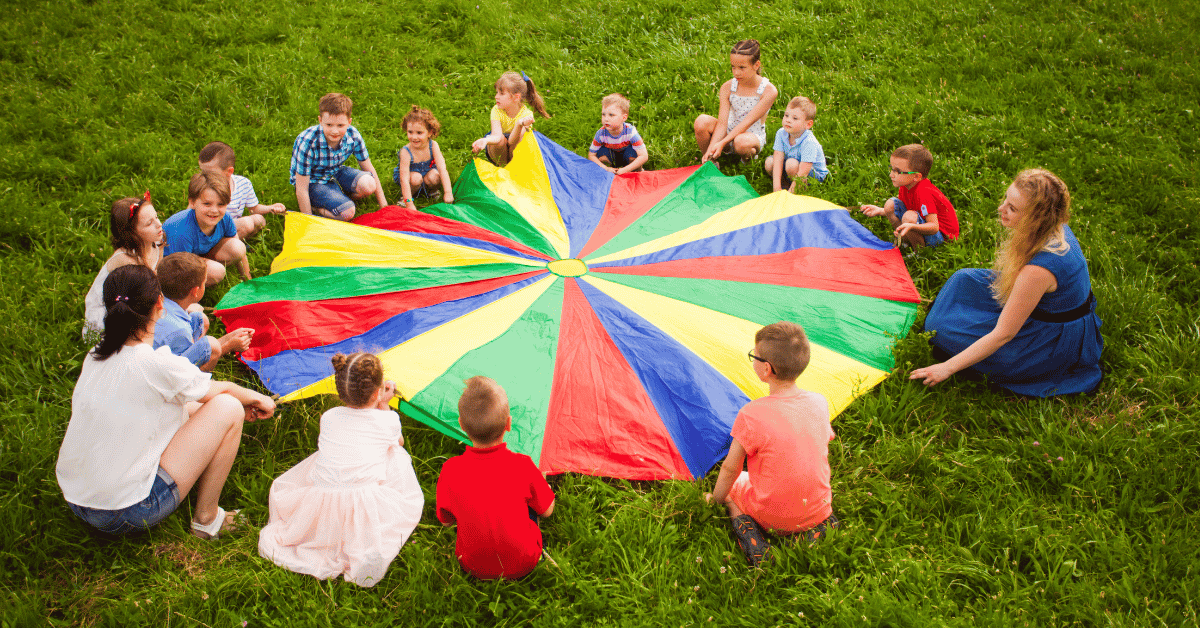 A group of kids and adults on the grass all holding onto a rainbow parachute to play a game representing a metaphor for how we uphold limiting societal beliefs