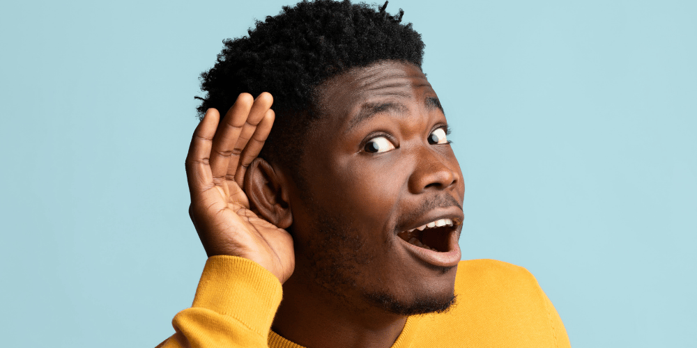 A man wearing a. yellow shirt with a blue background holding his hand around his ear with a very curious excited expression reflecting learning about the truth about ascension symptoms.