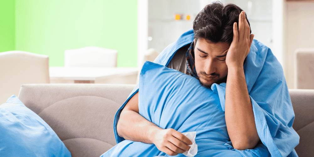 man sitting on a sofa with a blue blanket over his shoulders and holding a blue pillow with this hand on his head looking unwell as a reflection of ascension symptoms or actual illness. 