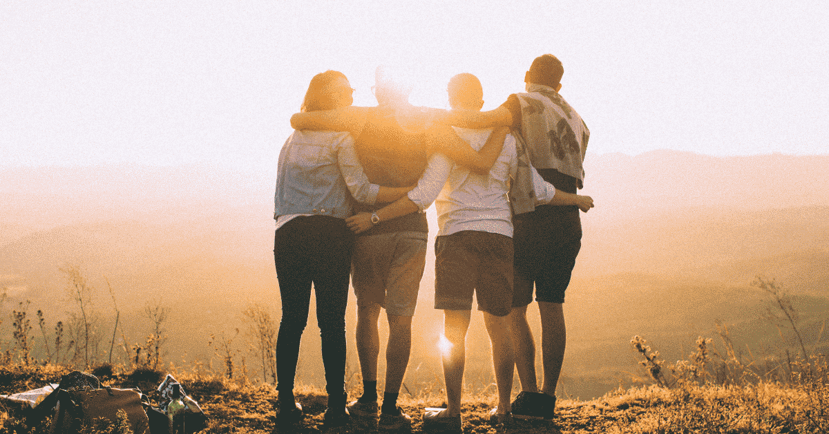 Four people with their arms all around one another with the view from their backs with the sun setting in front of them reflecting what it means to be free